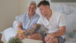 © luciano - A 70-year-old woman and a young 20-year-old grandson look at the boy's cell phone together while sitting on the couch at home. Friendship and affection in a multigenerational family