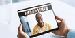 © Prostock-studio - A young doctor is engaged in an online consultation. He holds a tablet with a video call on the screen featuring a smiling patient. The setting is a bright room with a white table.