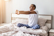 © Anastasiya - Young black man sitting in bed stretching arms forward after waking up, smiling and relaxed, representing healthy morning routine, flexibility and active lifestyle at home
