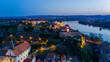 © AmazingAerialAgency - Aerial view of the illuminated Petrovaradin Fortress standing majestically above the Danube River, a beacon of history against the twilight sky, Petrovaradin, Vojvodina, Serbia.