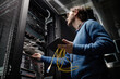 © Seventyfour - Middle aged Caucasian man working in server room holding tablet and yellow network cables, inspecting server rack equipment, focusing on data center maintenance and technology management