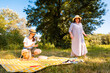 © _KUBE_ - Young Caucasian girl and her mother relaxing on a blanket in a green park enjoying family time and outdoor entertainment under sunny summer sky.