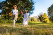 © _KUBE_ - Young Caucasian girl and her mother carrying a blanket through park greenery exploring a place for picnic enjoying summer holiday and family fun together. Low angle view
