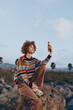 © SHOTPRIME STUDIO - Young woman with curly hair wearing rainbow sweater sits on rock outdoors smiling while taking selfie with smartphone in natural landscape during daytime with clear sky.