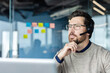 © Liubomir - Bearded call center agent with glasses and headset in a modern office, looking away thoughtfully while providing customer support and technical assistance with concentration and professionalism
