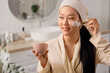 © Pixel-Shot - Beautiful young happy Asian woman with brush applying mask on her face in bathroom