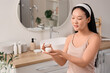 © Pixel-Shot - Beautiful young happy Asian woman with facial sheet mask and jar of cream in bathroom