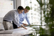 © fizkes - Collaborative data analysis. Two diverse multiethnic businessmen colleagues lean on office desk review printed charts graphs in marketing report discuss project results study statistical information
