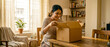 © CGI STOCKS - Smiling young South Asian woman happily opening a cardboard delivery box at a table in a cozy, sunlit home.