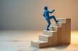© Maryna - Blue manikin ascends wooden stpic. Figure moves upward on stairs indicating progress and reaching a goal. Small person takes stpic on blocks for development and success.