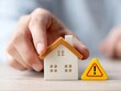 © Jumpei - 住宅の模型に手を添えて注意標識を横に並べる家づくりや不動産のリスクを懸念する男性