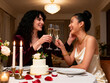 © Abhi - Two women celebrating their anniversary at a beautifully set intimate dining table at home. The table has a small two-tier cake with white frosting and fresh flowers on it, two tall champagne flutes b