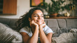 © Kowit - A young woman with curly hair smiles thoughtfully while resting her chin on her hands, sitting comfortably on a couch in a cozy, plant-filled living room.