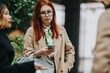 © qunica.com - Red-haired business worker checks her smartphone while standing with colleagues outdoors. A small group of business workers converse casually during a brief street-side meeting.