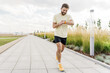© muse studio - Bearded man running on a wide paved park path, checking his fitness data on a bright smartwatch under a cloudy sky