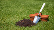 © Magdalena Wojaczek - A bright silver garden trowel, a small mound of dark fertile soil, and three empty terracotta pots are perfectly placed on lush green grass for spring planting.