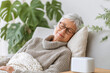 © kora studio - Aging woman relaxing on sofa, wearing glasses, with cozy sweater and knitted blanket. She appears content and peaceful, surrounded by indoor plants