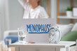 © Pixel-Shot - Paper with word MENOPAUSE, alarm clock, glass of water and pills on table against woman at home, closeup