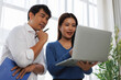 © Tj - Two Asian business colleagues, a man and a woman, standing and engaging in a focused discussion, collaborating on a project by looking at a laptop computer in a bright office environment