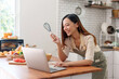 © Crystal - Smiling woman wearing an apron consulting a digital recipe on a laptop while holding a whisk in a modern kitchen