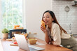 © Crystal - Young woman smiling, holding a croissant and leaning on a kitchen counter with a laptop and fresh food