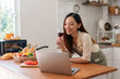 © Crystal - Woman smiling and holding a glass of wine, videoconferencing on a laptop while preparing food in a modern kitchen