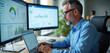 © miss irine - Mature man in blue shirt checks charts on computer screens. He works in office analyzing business data on laptop and desktop monitors. Uses CRM software for sales.