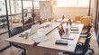 © zyla - Vibrant collaborative workspace with multiple laptops and documents on a large wooden table in a sunlit office.
