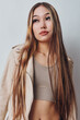 © SHOTPRIME STUDIO - Young woman with long hair wearing beige jacket and crop top standing against white background looking away with serious expression and natural makeup.