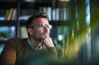 © EricMiguel - Thoughtful executive in a brown sweater and round glasses resting chin on hand, gazing out a bright corner office window past green plants