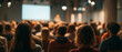 © perfectlab - Audience attending a conference or seminar in a dimly lit room with a speaker presenting on stage and a large screen in the background