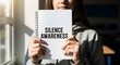 © Afif - A woman holds up a notebook with silence awareness text in a room.