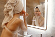 © Vlad - Beautiful smiling young woman in lingerie with towel on head, making face massage using roller, standing in bathroom, looking at mirror. Beauty facial procedures