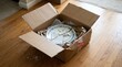 © David - Broken ceramic plate inside open cardboard box with packing materials, indicating damaged delivery on a wooden floor.