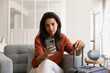 © fizkes - Young focused woman using smartphone while holding suitcase handle seated in cozy armchair at home or hotel room. Mobile application usage for accommodation booking, tickets buying, vacation planning