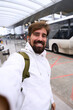 © CarlosBarquero - Vertical. Smiling caucasian millennial man taking a selfie outside an airport terminal with a shuttle bus behind, wearing hoodie, backpack and earbuds. Bright canopy architecture, travel and movement.