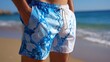 © Harry - A man wearing blue and white swim trunks stands on a sandy beach with the ocean in the background.