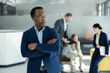 © Dejan - Young african entrepreneur with crossed arms posing confidently in a corporate setting with diverse colleagues in the background