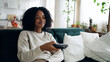 © DMegias - Young black woman comfortably sitting on a sofa, holding a remote control, enjoying television entertainment and leisure time