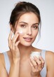 © YOUSSEF KETTOU - Portrait of a young woman applying moisturizer to her face, holding a jar of cream in her hand, with soft studio lighting