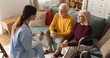 © Stockphotodirectors - Nurse sits on the floor and talks to an elderly couple on the couch in their living room. The couple listens and engages with the nurse during the home visit.