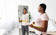 © Prostock-studio - Overweight young black woman measuring her waist with tape near mirror at home, satisfied with slimming result, free space. Plus size African American lady being successful in weight loss