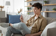 © Prostock-studio - Cheerful Young Asian Man Using Smartphone Texting Online And Scrolling News Feed In Social Media Application Sitting On Couch At Home. Happy Guy Reading Message On Cell Phone. New Mobile App Concept