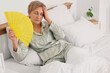 © Pixel-Shot - Sweaty mature woman with fan experiencing menopause in bedroom