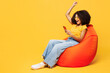 © ViDi Studio - Full body young woman of African American ethnicity wear t-shirt casual clothes sit in bag chair use mobile cell phone do winner gesture isolated on plain yellow orange background. Lifestyle concept.