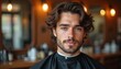 © Pete - Young man with wavy hair and beard sits in barber chair wearing cape. He looks directly at camera with soft gaze. Blurry barber shop background has warm lighting.
