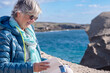 © luciano - A smiling mature woman sits on a rocky beach, holding a book and relaxing while reading. Vacation, retirement, and the blue waters of the ocean.