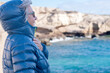 © luciano - Happy elderly lady standing on a rocky beach smiling, gazing at the cliffs while covering herself with a jacket against the cold wind. Vacation, retirement leisure, and the blue waters of the ocean
