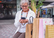 © luciano - Smiling elderly woman sitting in a shopping mall after shopping for black friday while using mobile phone. Attractive senior lady with shopping bags, consumerism concept