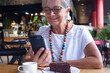 © luciano - Attractive senior woman relaxing for a break at coffee shop in mall using mobile phone enjoying a coffee cup and a slice of cake. Carefree smiling elderly woman with eyeglasses and necklace